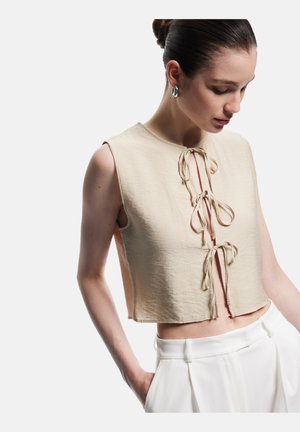 Top beige tagliato con scollatura rotonda e dettaglio di nodo frontale. Realizzato in un tessuto testurizzato, presenta un taglio senza maniche e una vestibilità ampia.