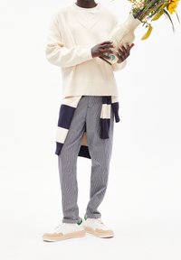 Maglione beige a scollo a V, pantaloni a righe blu e bianchi, e sneakers bianche con accenti beige e verdi. Tienendo un vaso decorato con fiori.