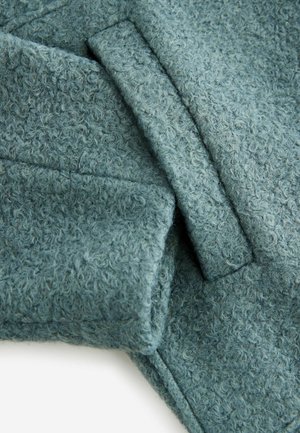 Texturiertes, weiches, türkisfarbenes Gewebe mit einer flauschigen Oberfläche, das gefaltete Kanten und Nähte aufweist und so ein geschichtetes Aussehen erzeugt.