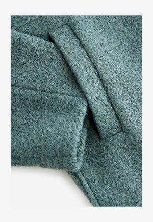 Teksturirana, mehka, teal tkanina z puhastim zaključkom, ki ima pregibne robove in šive, kar ustvarja plastičen videz.