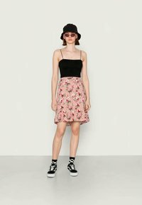 Mulher a usar um chapéu preto tipo balde, óculos de sol cor-de-rosa, top preto de alças finas, saia floral cor-de-rosa, meias às quadrados e sapatilhas pretas, em pé contra um fundo simples.