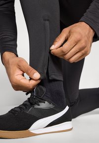 Melnas sporta legingi ar sānu rāvējslēdzējiem, cieši pieguļoši, apvienoti ar melnām un baltām kedām. Gluda tekstūra, minimāls dizains.