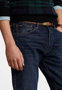 Man som bär mörkblå denimjeans med brunt bälte och en svart-grön rutig tröja, med handen vilande vid sidan, stående mot vit bakgrund.