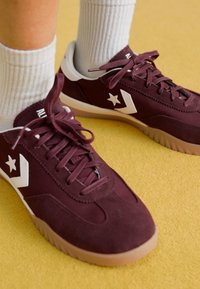 Burgundy velúr sportcipők fehér csillag és chevron díszítéssel, lapos fűzőkkel és texturált gumi talppal, egy sárga felületen pihenve.