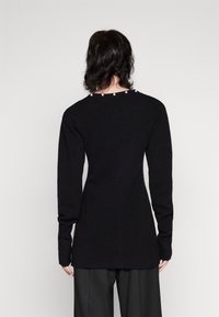 Persona con cabello oscuro hasta los hombros, vistiendo una camiseta negra de manga larga ajustada con detalles de perlas en el escote trasero y pantalones negros.