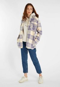 Geruite fleecjas in crème en paars gedragen over een lichte hoodie. Heeft zakken, een ontspannen pasvorm en blauwe jeans met een cropped zoom.