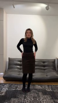 Woman with long hair wearing a black long-sleeve top, dark skirt, and tights stands barefoot on a patterned rug in front of a black leather couch.