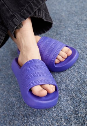 Pieds portant des sandales slides violettes avec des brides texturées, reposant sur un sol moquetté bleu, avec un pantalon noir partiellement visible.