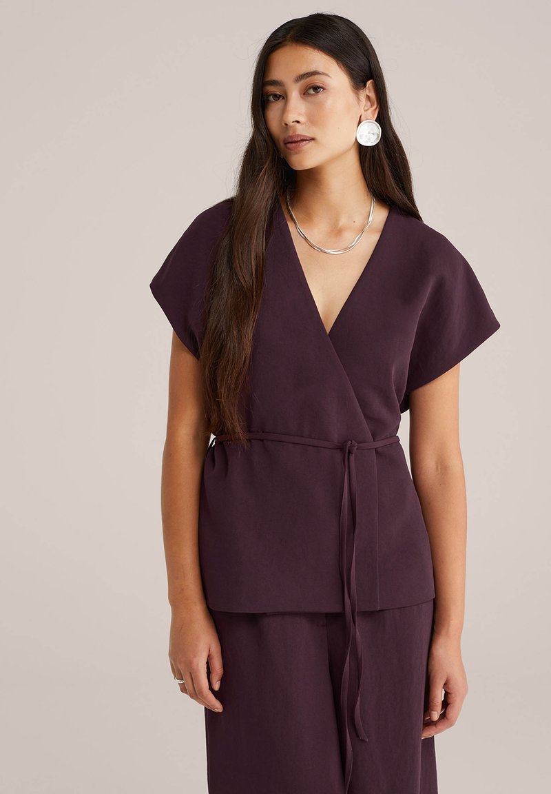 Donna con lunghi capelli scuri che indossa un top avvolto di un profondo viola e pantaloni abbinati, collana d'argento e grandi orecchini rotondi, sfondo neutro.