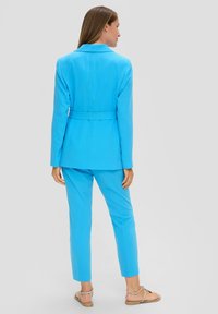 Blazer ajusté bleu clair avec un pantalon slim assorti. Il présente une ceinture à la taille et un tissu lisse. Le modèle porte des sandales plates.