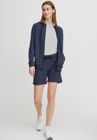 Jūras spēku džemperis ar kapuci, pelēks T-krekls un atbilstošas šortus, visi izgatavoti no mīksta auduma. Baltas sporta kurpes ar pelēkiem akcentiem.