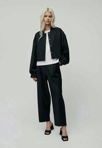 Svart kort bomberjacka med tryckknappar, tillsammans med en vit topp och svarta vida byxor. Har en slät textur och en avslappnad passform.