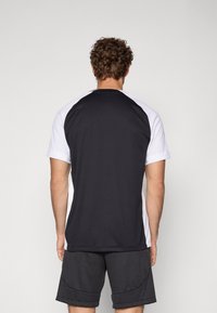 Tricou sportiv alb-negru cu mâneci scurte, având un design raglan și o textură fină. Vedere din spate, purtat cu pantaloni scurți gri.