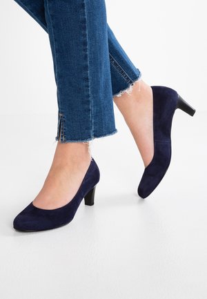Fødder iført mørkeblå ruskind midhælspumps kombineret med cropped blå denimjeans med flossede kanter og sidelommer.