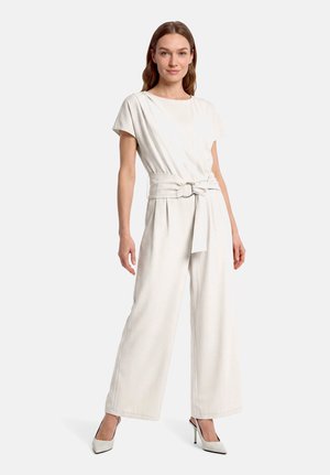 Kvinde står iført en off-white kortærmet jumpsuit med bredt bælte og højhælede spidse sko på en hvid baggrund.
