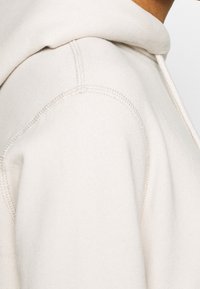 Hellbeiger Kapuzenpullover aus einem weichen, strukturierten Stoff mit glatter Oberfläche, sichtbaren Nähten und Kapuze mit Kordelzug.