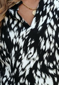 Zwart-witte blouse met patroon en een V-hals. De stof heeft een gladde textuur met contrasterende penseelstrekenpatronen. Gouden kettingaccent.