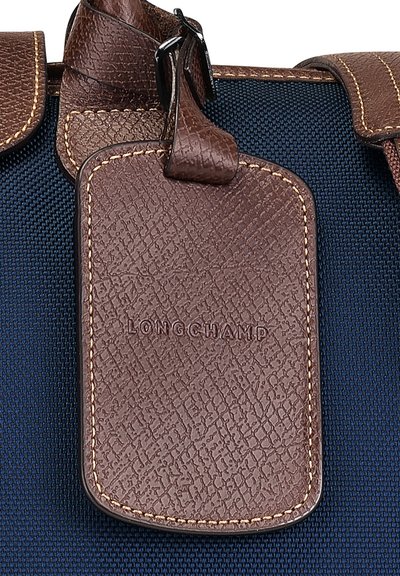 Brauner Ledergepäckanhänger mit strukturiertem Oberflächen, versehen mit geprägtem "LONGCHAMP"-Logo, angebracht auf einem marineblauen Stoffhintergrund.