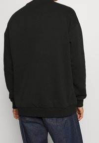 Persona vistiendo un sudadera negra básica y jeans azules, mostrada de espaldas contra un fondo claro y liso.