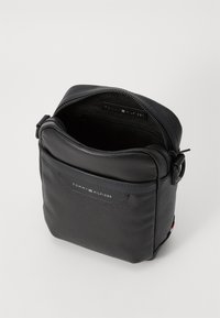 Bolso cruzado de cuero negro con cierre en la parte superior, acabado texturizado, un bolsillo frontal y una etiqueta de marca. Forma rectangular compacta.