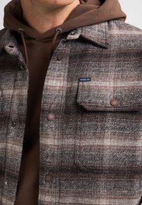 Bruin en grijs geruite shirtjack gemaakt van textuurstof, met daaronder een bruine hoodie, knoopdetails en een borstzak.