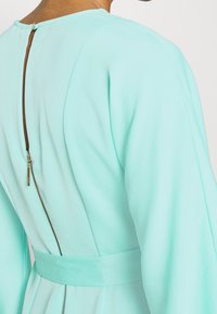 En mintgrön klänning med en slät tyg, långa ärmar, en figursydd midja med bälte och en synlig dragkedja bak med en dragflik.