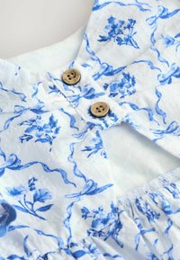 Vestito di cotone azzurro con motivi floreali e a nastro, caratterizzato da due bottoni di legno sul davanti e dettagli arricciati in vita.