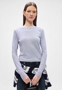 Λαμπερό μωβ πουκάμισο με μακριά μανίκια και στρογγυλή λαιμόκοψη, διακοσμημένο με κεντημένο λογότυπο. Συνδυάζεται με σκούρα τζιν και ένα μπλε και άσπρο πλεκτό κασκόλ.