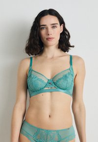 Turkio spalvos nėriniuotė liemenėlė su permatomi tinkleliais, gėlių raštais, reguliuojamomis petnešėlėmis ir dekoratyvine juostele centre.