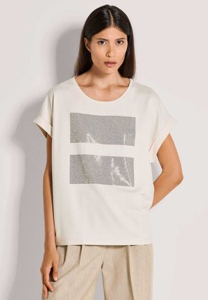 Hellbeige T-Shirt mit einem großen silbernen Paillettenquadrat-Design und dem Text "THIS IS AMAZING" in der Mitte; locker geschnitten mit hochgekrempelten Ärmeln.