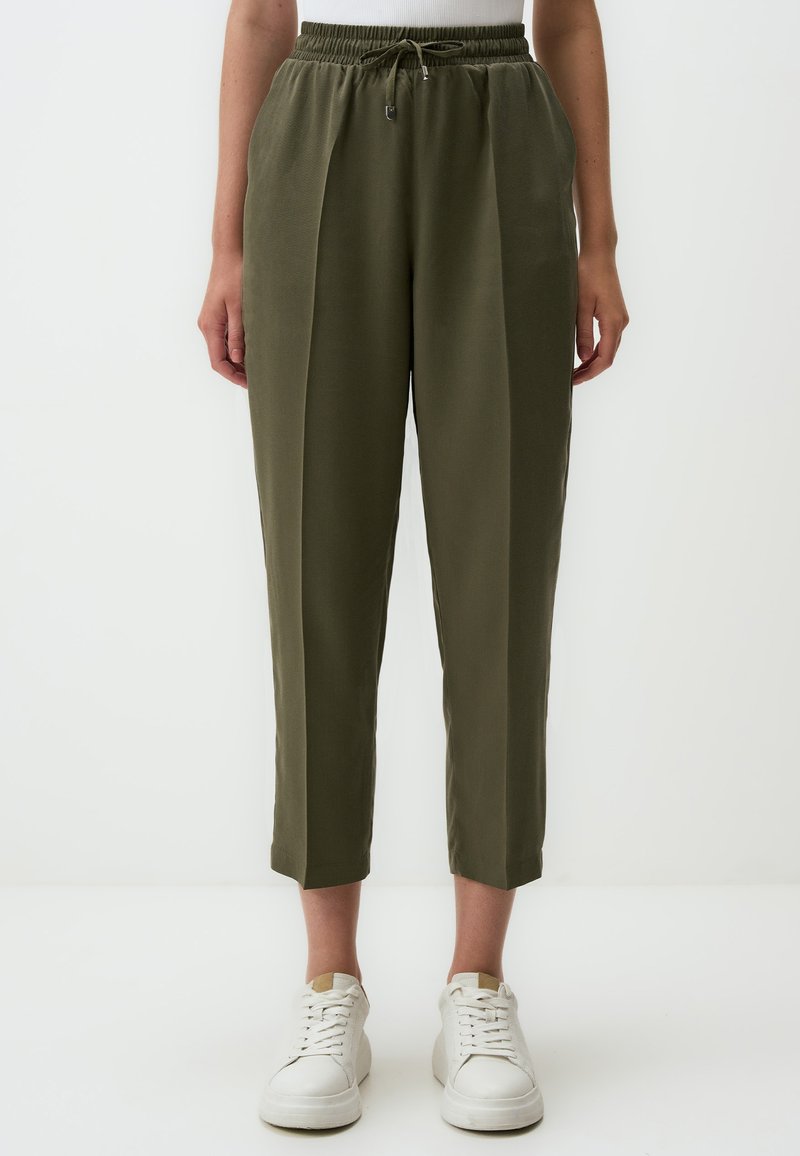 Pantalons verts olive, fuselés avec une taille élastique, cordon de serrage et plis latéraux. Texture lisse, longueur cropped, associés à des baskets blanches.