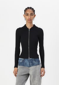 Svart ribbet hettegenser med glidelås, lange ermer og et figursydde design. Kombinert med lyseblå og grå lagdelte bukser med en rynket tekstur.