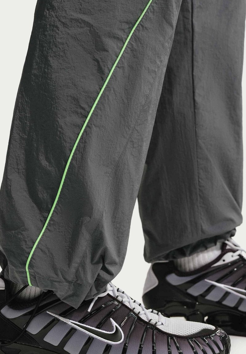 Persoon draagt donkergrijze broek met een groene streep en zwart-witte Nike-sneakers, broek bij de enkels bijeen, staand op een lichte achtergrond.