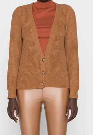 Persoon die een getextureerde bruine gebreide cardigan met knopen draagt over een roestkleurige coltrui en beige neplederen broek, staand tegen een effen achtergrond.