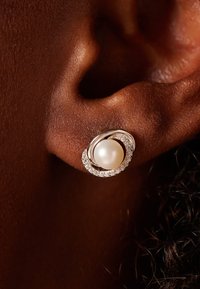 Boucles d'oreilles en argent avec une perle blanche ronde au centre, entourée d'un bandeau ondulé et de petites pierres précieuses claires, mises en valeur sur une peau sombre.