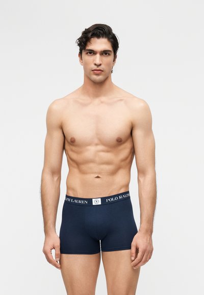 Bărbat musculos cu păr închis la culoare, purtând boxer briefs Polo Ralph Lauren albastru închis, pe un fundal simplu, deschis la culoare, privind înainte cu o expresie neutră.