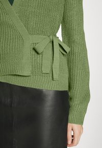 Grüne gestrickte Strickjacke mit Wickeldesign, mit einem Bindeband in der Taille, strukturiertem Stoff und gerippten Bündchen, kombiniert mit einem schwarzen Lederrock.