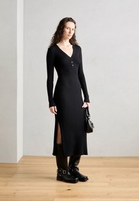 Black ribbed knit dress with long sleeves, a deep V-neck, a buttoned placket, and side slits. Paired with black knee-high boots and a handbag.