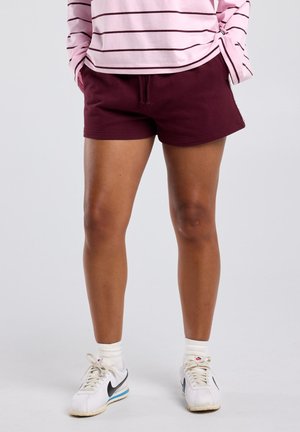 Persoon draagt bordeauxrode shorts, roze gestreepte top met lange mouwen, witte sokken en witte Nike sneakers, staat met handen in de zakken.