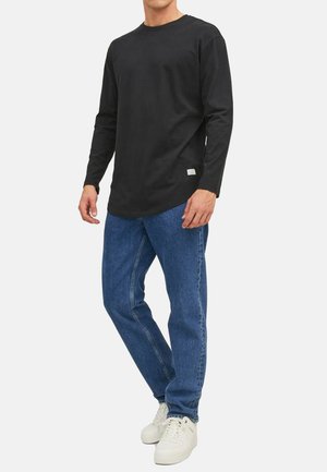 Sort langærmet skjorte lavet af bomuld, parret med blå jeans og hvide sneakers. Skjorten har en rund nederkant og et diskret logo-tag.