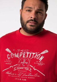 T-shirt rossa in cotone con stampa grafica bianca che presenta immagini di canottaggio e testo. Collo rotondo e maniche corte.