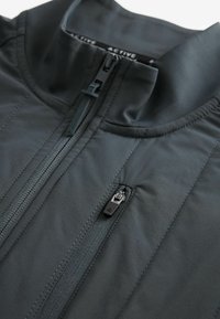 Chaqueta de cremallera gris oscuro con cuello alto, con tejido suave y detalles de costura en contraste. Incluye dos bolsillos con cremallera.