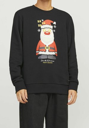 Une personne porte un sweat-shirt noir arborant un père Noël en dessin animé avec un nez rouge, du texte festif et des flocons de neige, sur fond uni.