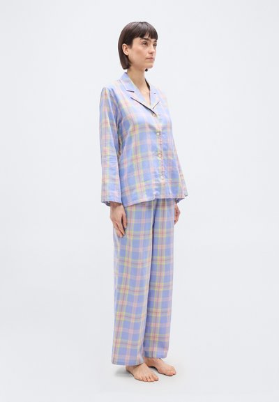 Γυναίκα που στέκεται ξυπόλητη, φοράει πιτζάμα με παστέλ καρό πουκάμισο με κουμπιά και ταιριαστό παντελόνι πιτζάμα με φαρδιά πόδια, μπροστά σε απλό φόντο.
