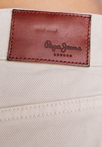 Bežna tkanina s dijagonalnim tkanjem, koja sadrži smeđi kožni pečat utisnut s natpisima "Pepe Jeans" i "LONDON", prošiven za izdržljivost.