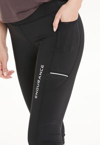 Musta urheilullinen legginssit sileällä pinnalla, joissa on sivutasku ja valkoinen "ENDURANCE"-logo, suunniteltu mukavuutta ja suorituskykyä varten.