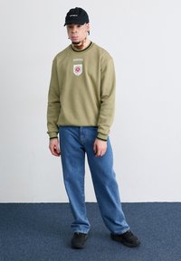 Olíva zöld Adidas pulóver logóval, kék farmernadrággal párosítva. Fekete sapka és fekete cipő egészíti ki az összképet. Egyszerű, laza stílus.