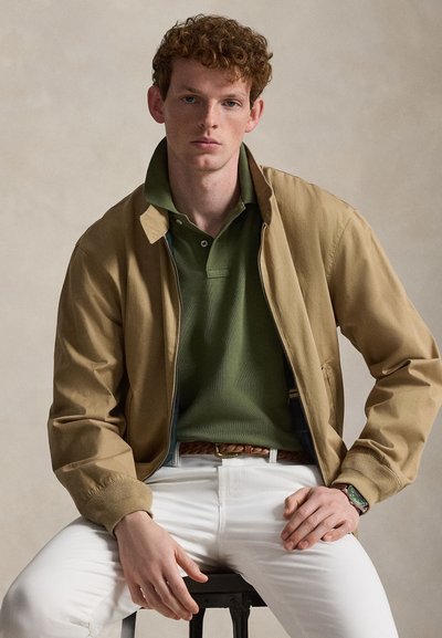 Väriltään tan bomber-takki vihreän poolopaidan päällä, yhdistettynä valkoisiin housuihin. Takissa on resorit hihansuissa ja ruskea kudottu vyö. Ranteessa vihreällä kellotaululla varustettu rannekello.