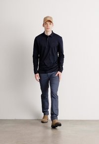 Langärmliges, dunkelblaues Polo, graue Hose und beiger Hut. Das Modell geht auf einem hellen Betonboden mit einem neutralen Hintergrund.