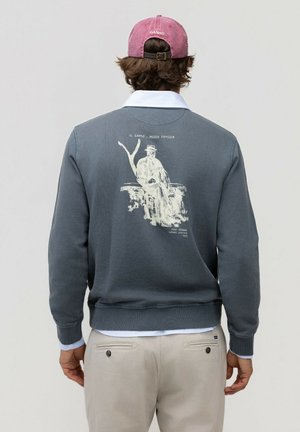 Hombre con sudadera gris con cuello blanco y arte vintage impreso en la espalda, pantalones beige y gorra rosa, de espaldas.